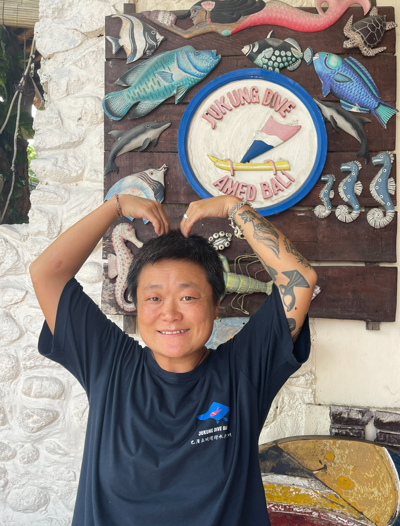 Smiling Jukung Dive Bali team member standing in front of the Jukung Dive logo sign at the dive resort in Bali